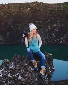 a woman sitting on top of a large rock next to a lake with a camera