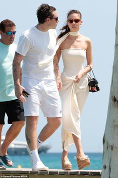 a man and woman walking on a dock next to the ocean while wearing white clothing