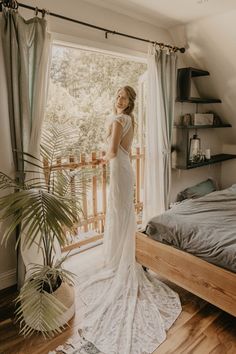 a woman standing in front of a bed wearing a wedding dress and looking out the window