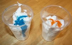 two plastic cups filled with whipped cream and blue and orange icing on top of a wooden table