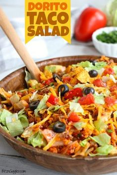 a wooden bowl filled with taco salad next to lettuce, tomatoes and black olives