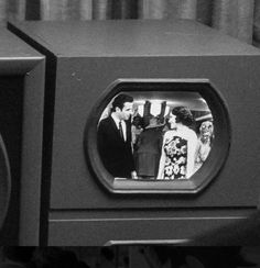 a black and white photo of two people in front of an old tv with the image of a man and woman on it