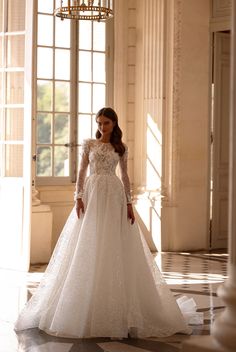 a woman in a white wedding dress standing by a chandelier and window with her hands on her hips