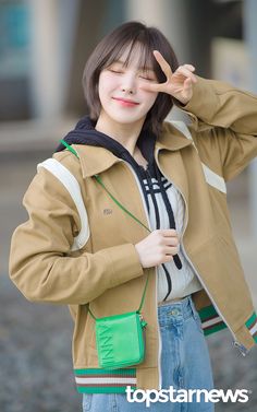 a young woman wearing a tan jacket and green purse posing for the camera with her hand up to her face