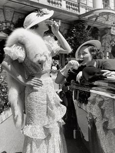 a woman in a dress and hat standing next to a man holding a stuffed animal