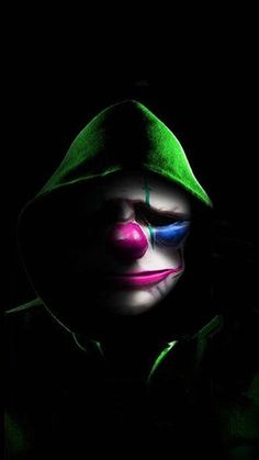 a creepy clown in the dark with his face painted pink and blue, wearing a green hoodie