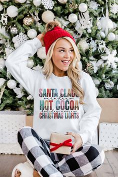 a woman sitting in front of a christmas tree wearing a santa clause sweatshirt and plaid pants