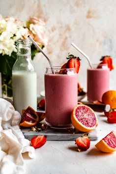 two glasses filled with pink smoothie next to sliced oranges and strawberries on a table