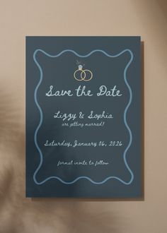 a blue save the date card with two wedding rings on it next to a plant