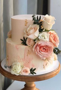 a three tiered wedding cake with pink and white flowers on the top, sitting on a wooden stand