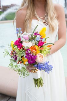 a woman in a white dress holding a bouquet of colorful flowers and greenery near a fountain