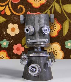 a metal robot with eyes and nose sitting in front of a flowery wallpaper