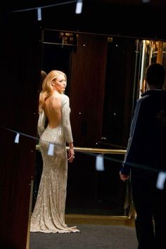 a woman standing in front of a mirror with her back turned to the camera and wearing a long sleeved dress