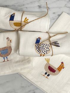 three napkins with embroidered chickens on them sitting on a marble counter top next to each other