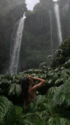 a woman standing in front of a waterfall surrounded by lush green foliage