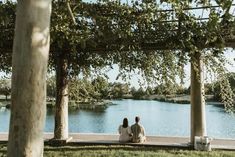 two people sitting on a bench under some trees by the water and looking at something in the distance