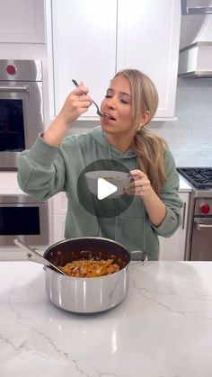 a woman in a kitchen holding a spoon over a pan filled with food and looking at the camera