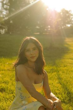 a woman in a yellow dress sitting on the grass with her legs crossed and looking at the camera