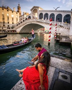 a man and woman sitting on the edge of a pier next to a canal with gondola passing by