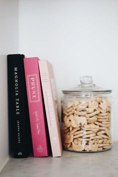 some books are sitting on a shelf next to a glass jar filled with nuts and crackers