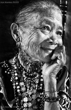 an old woman with necklaces on her neck and hand to her chin, looking at the camera