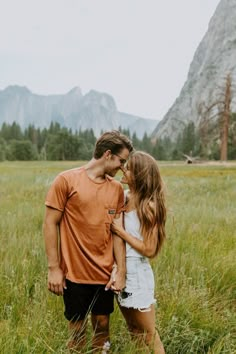 a man and woman standing in tall grass with mountains in the background on a cloudy day