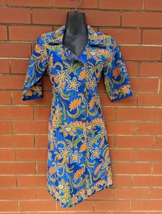 Found! 🔍 Super cool shirt-dress with asymmetrical button line and ability to be buttoned high necked or folded open. We especially love the cobalt blue colour contrasted with the orange tropical paisley floral swirling all over. 📋 - This dress is in excellent vintage condition with no noticeable defects. Made by M3rlin.  Size S.  👗 Measurements (taken flat):  - 93cm length  - 40cm shoulder  - 44cm bust  - 38.5cm waist  - 35.5cm sleeve length  - 15cm cuff width ------------------------------ Retro Blue Button-up Dress, Blue Collared Shirt Dress With Placket, Blue Shirt Dress With Placket For Work, Blue Knee-length Shirt Dress With Placket, Blue Cotton Button-up Dress, Vintage Blue Button-up Dress, Blue Button Cuffs Dress For Work, Blue Cotton Shirt Dress With Button Closure, Blue Cotton Formal Dress