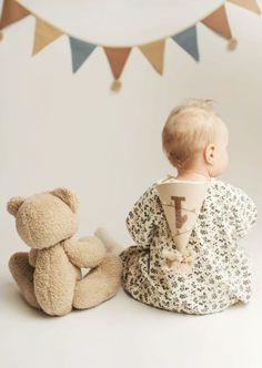 a baby sitting next to a teddy bear on the floor with bunting flags in the background