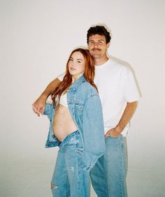 a man and woman standing next to each other in front of a white wall wearing jeans