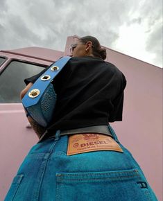 a woman wearing a blue purse standing next to a pink car with clouds in the background