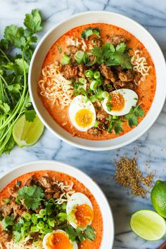 two white bowls filled with soup and topped with hard boiled eggs, cilantro, green onions, lime wedges