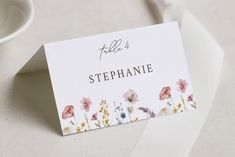 a place card with watercolor flowers on it next to a white cup and saucer