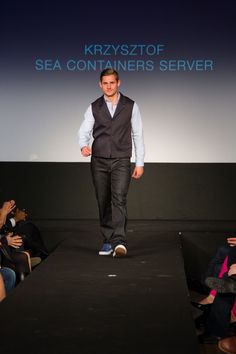 a man is walking down the runway wearing a vest and jeans, while people watch