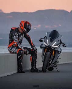 a person sitting on a ledge next to a motorbike and helmet, with mountains in the background