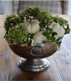 a metal bowl filled with white pumpkins and greenery on top of a wooden table