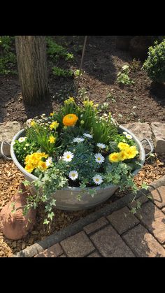 a planter filled with lots of flowers on top of a brick walkway next to a tree