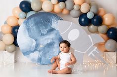 a baby sitting on the floor in front of balloons and a rocket ship wall decoration