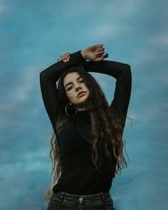 a woman with long hair standing in front of a blue sky and holding her hands behind her head