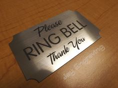 a metal name tag that says please ring bell thank you on a wooden table top