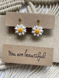 two small white and yellow daisy earrings on top of a brown card with the words you are beautiful