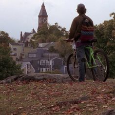 a man standing next to his bike on top of a leaf covered hill with houses in the background