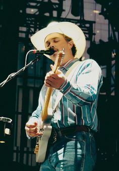 a man wearing a cowboy hat and holding a guitar in front of a microphone on stage