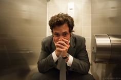 a man in a suit and tie sitting on a urinal with his hands clasped to his face