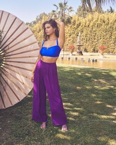 a woman is standing in the grass with an umbrella and wearing purple wide legged pants