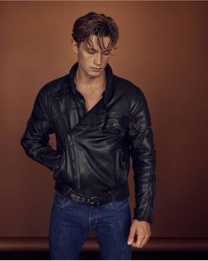 a man wearing a black leather jacket and jeans standing in front of a brown wall