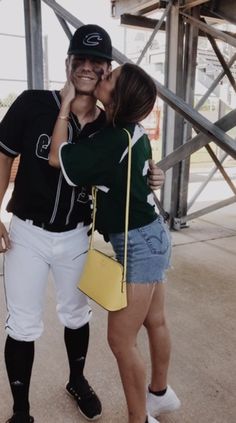 a couple of people that are kissing each other in the dirt with one person wearing a baseball uniform and holding a yellow purse