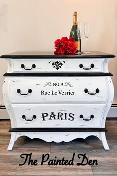 the painted dresser is white with black trim and has red flowers on top, next to a bottle of wine