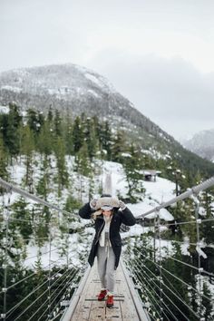 a woman walking across a suspension bridge in the middle of snow covered mountains and trees