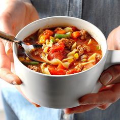 a person is holding a bowl of soup with noodles and meat in it, while the other hand holds a spoon