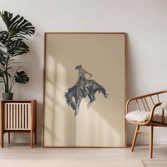 a framed photograph of a cowboy on a horse in a room next to a chair and potted plant